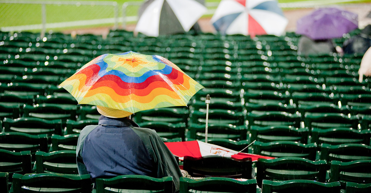 A person sits alone under a colorful umbrella in the stands of a baseball field.