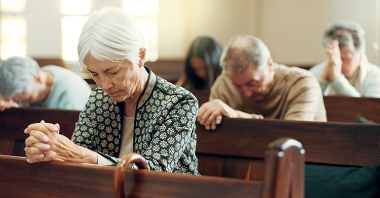 A white-haired woman sits in a pew and prays. Others pray in pews behind her