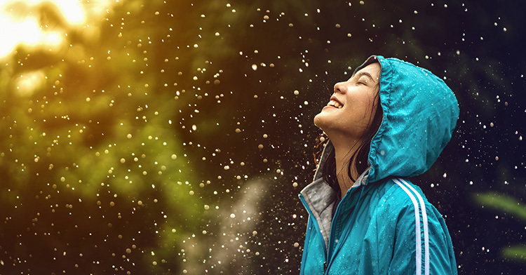 A woman wearing a blue rain jacket looks up at falling rain against a golden sky and smiles