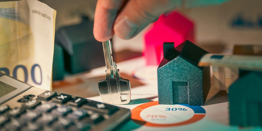 Image of a person holding a key in the midst of small models of houses, money, and a calculator