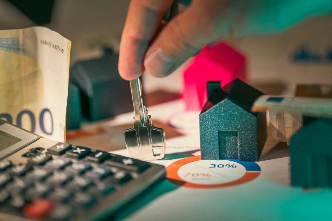 Image of a person holding a key in the midst of small models of houses, money, and a calculator